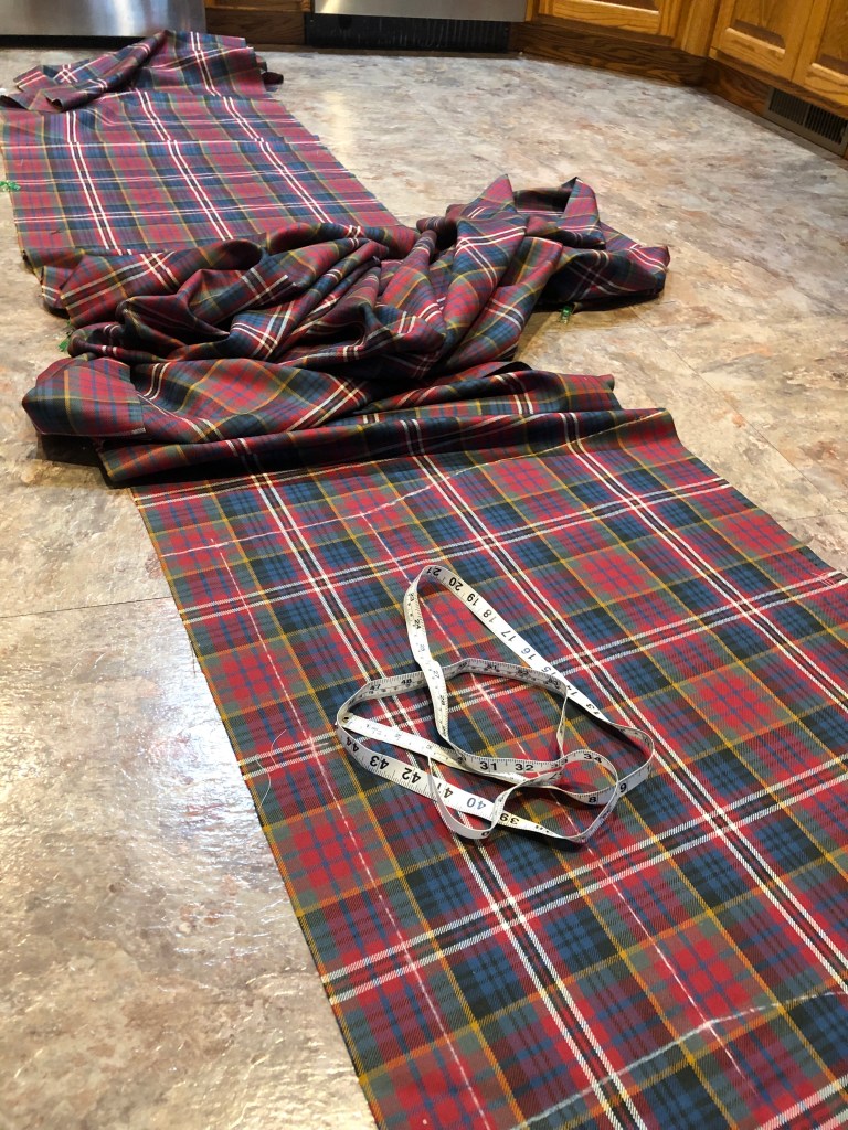 MacPherson Red Muted tartan spread across kitchen floor with most of it piled up in the middle, long tape measure on the kilt apron, which is marked in chalk
